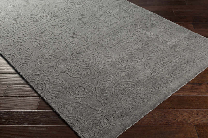 Close-up of charcoal Eversholt Clearance Rug showing floral, hand-loomed design and wool texture on dark wood floor. Corner
