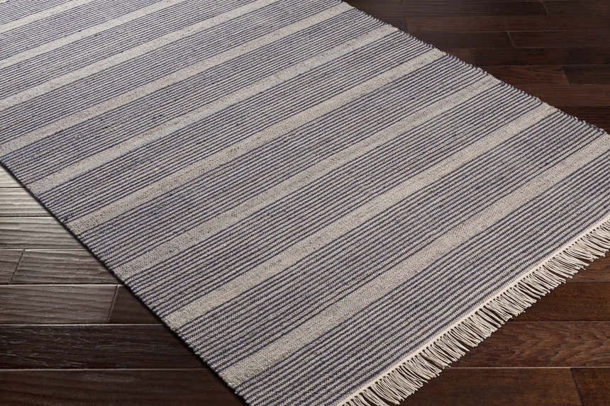 Close-up of the Gambalidio Area Rug. Navy and off-white striped, hand-woven wool-jute rug with fringe on dark wood floors. Corner