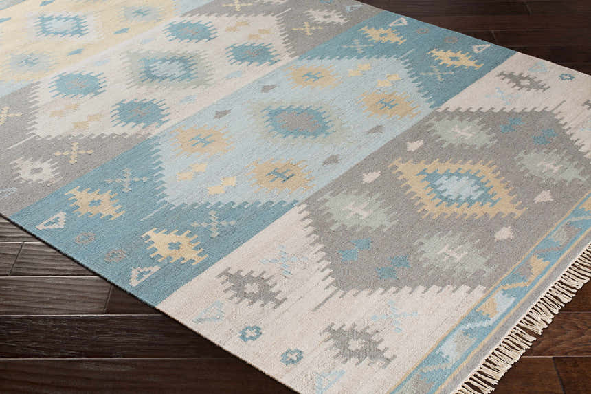 Close-up of hand woven Heth Carpet. Features a geometric pattern in beige, blue, charcoal, and tan on dark wood floors. Corner