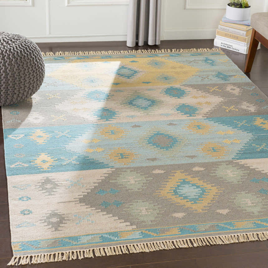 Living room scene with a Heth Carpet, gray pouf, and books. The rug has beige, blue, and charcoal colors. Roomscene-Rectangle