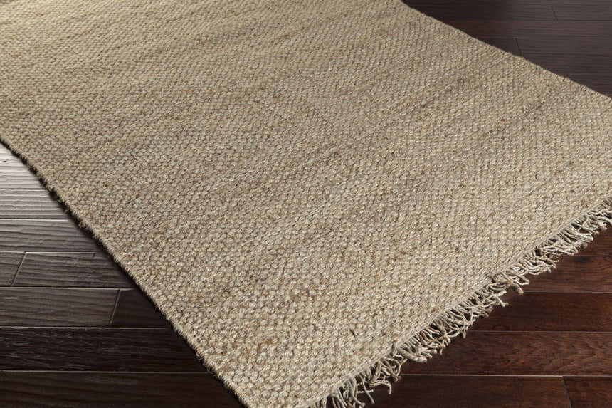 Close-up of Hovland Hand Woven Natural Jute Rag Rug showing natural jute color and fringe detail on dark hardwood floor. Swatch