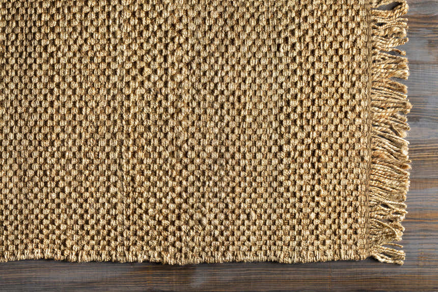 Close-up of the Hovland Hand Woven Natural Jute Rag Rug showing its texture, fringe, and tan color on a dark wood floor. Corner