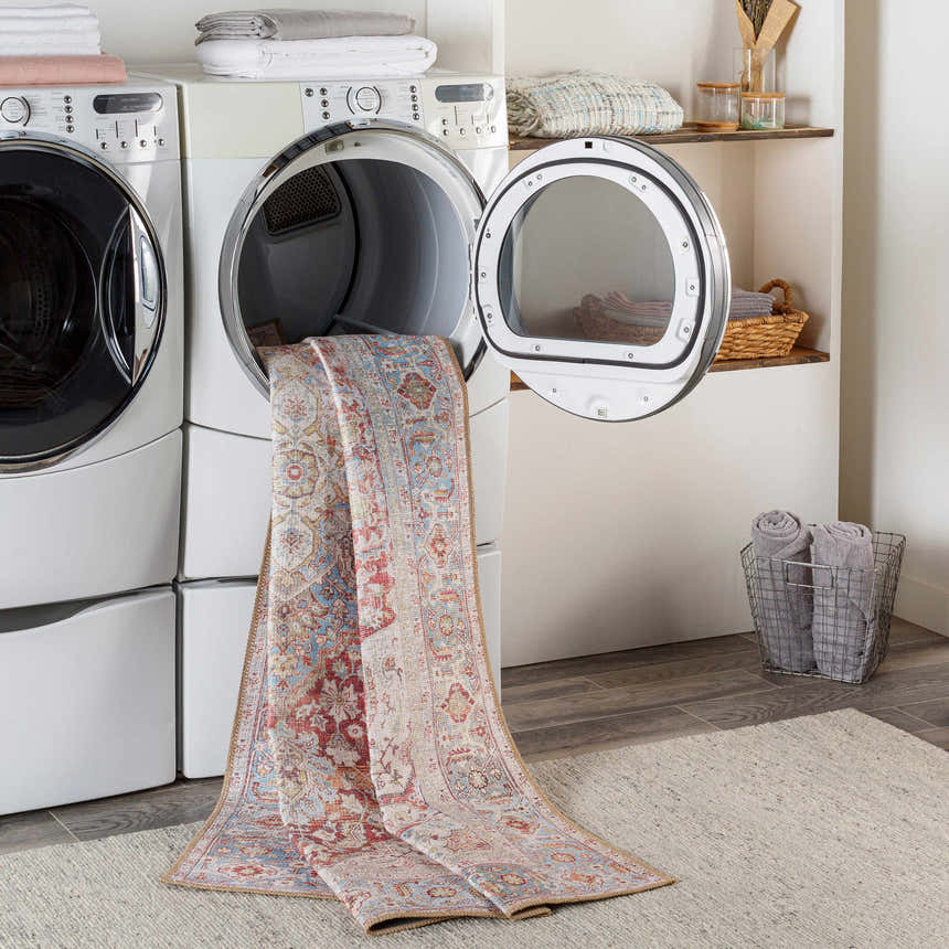 Laundry room scene with Cream Rust Leeston Washable Rug coming out of dryer. Bohemian vintage style rug in brick red, cream, and blue. Washing-Machine