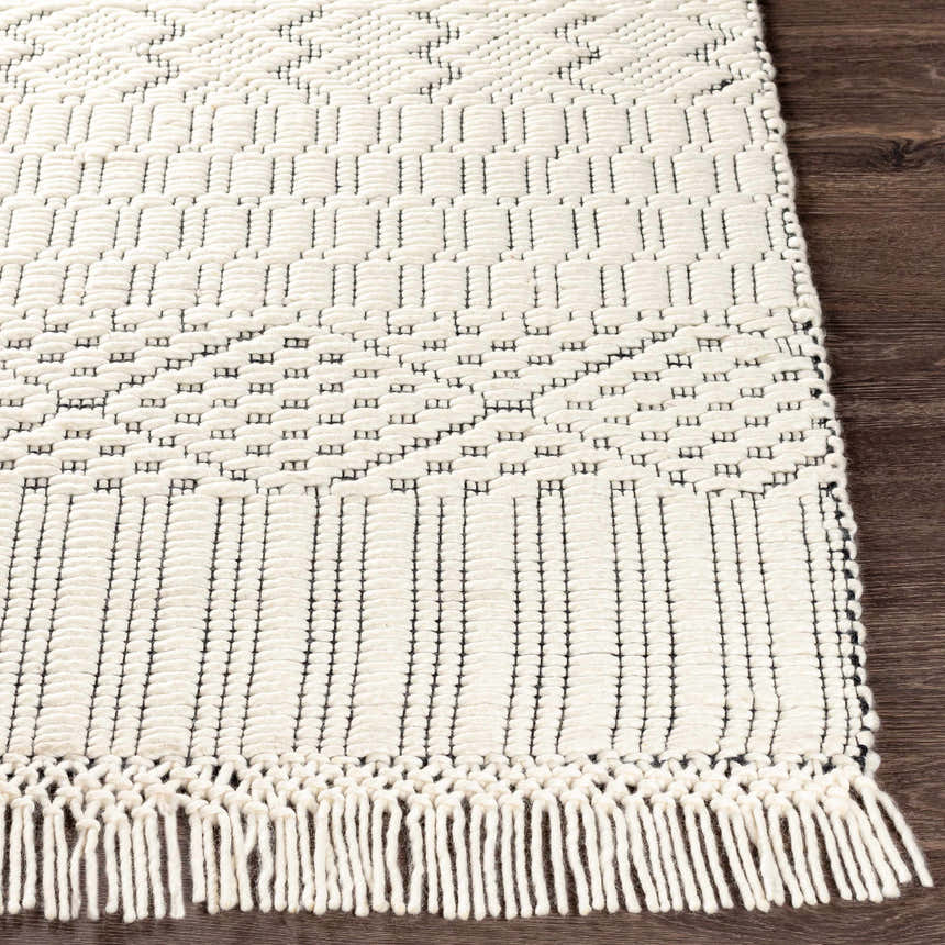 Close-up of the hand-woven Limoges Area Carpet. Cream wool rug with black geometric pattern and fringe on dark wood floor. Front