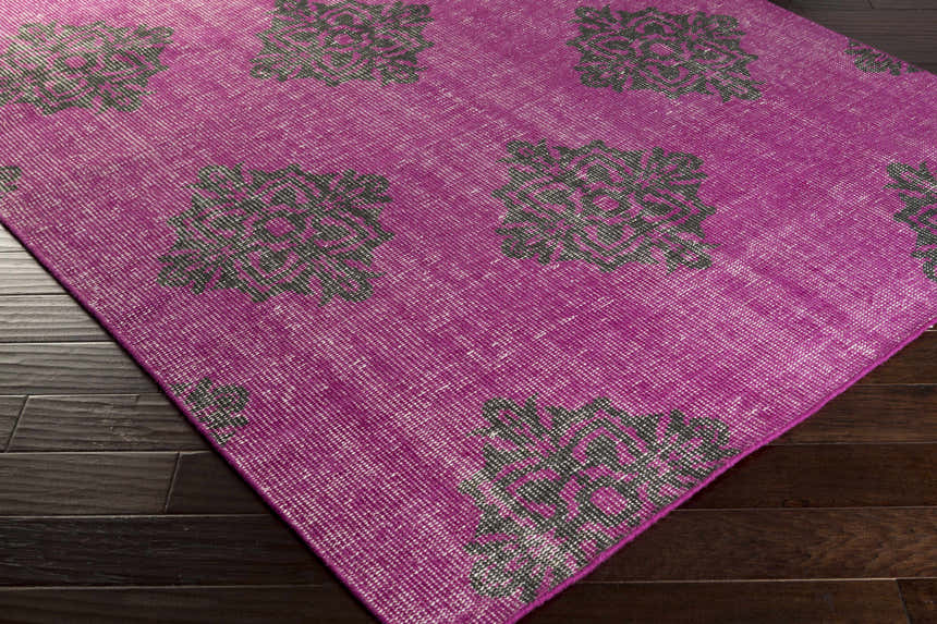 Close-up of the Mina Area Rug. Fuchsia wool rug with a traditional black floral pattern on dark wood flooring. Corner