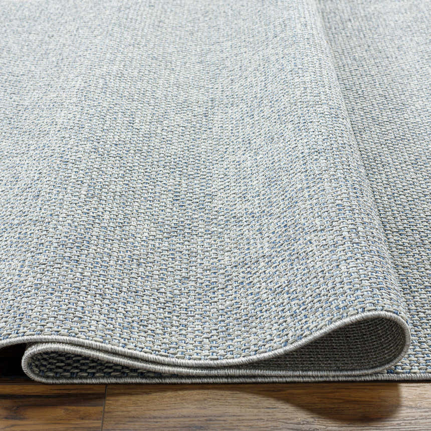 Close-up of the Mirit Area Rug showing its denim and light gray woven texture and folded edge on a wood floor. Texture