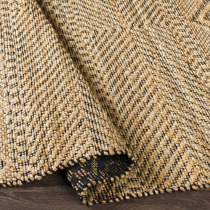 Close-up of the hand woven Nasugbu Jute Carpet. Tan jute rug with a subtle black geometric pattern and natural texture. Fold