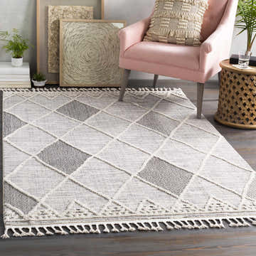 Living room scene with a Norvelt rug featuring a black, gray, and ivory diamond pattern and tassel trim. Pink chair and wood accents. Roomscene-Rectangle