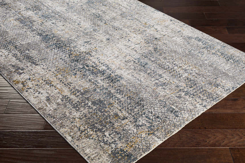 Close-up of the Odell Abstract Luxe Fringe Rug showing dark blue, gray, and mustard hues on dark wood flooring. Corner