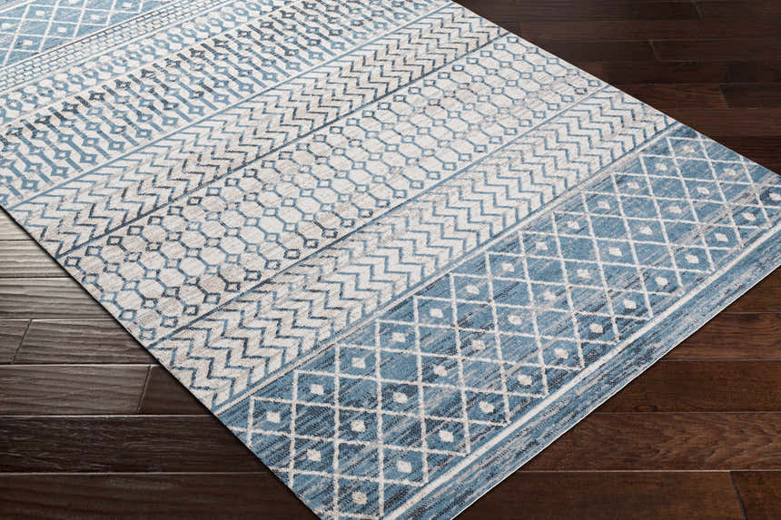 Close-up of the Pinamopoan Washable Area Rug. Features a geometric pattern in shades of blue, gray, and light beige. Corner