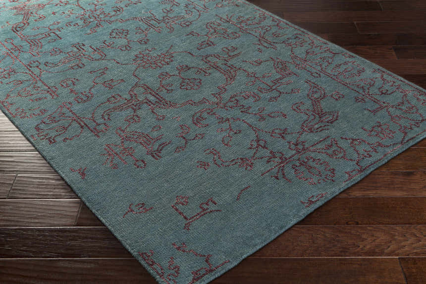 Close-up of the hand-knotted Redvale Area Rug. Teal rug with a burgundy, contemporary pattern on a dark wood floor. Corner