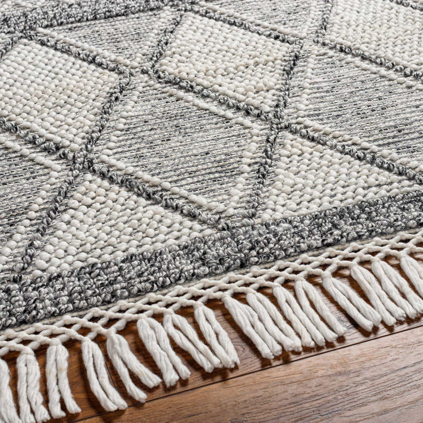 Close-up of the Rehn Area Rug, a hand-woven rug with a geometric diamond pattern and fringe, in dark gray, light gray, and taupe. Texture