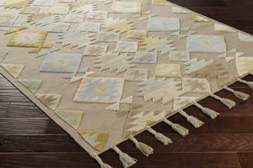 Close-up of Rippon Area Carpet. Hand-woven wool rug with beige, blue, tan, and yellow geometric pattern and tassels. Corner