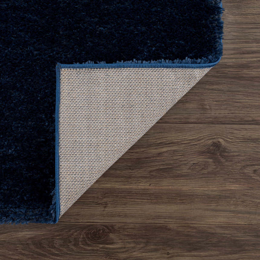 Close-up of the Navero Dark Blue rug showing its shag texture, backing, and navy blue edge on a dark wood floor. Fold