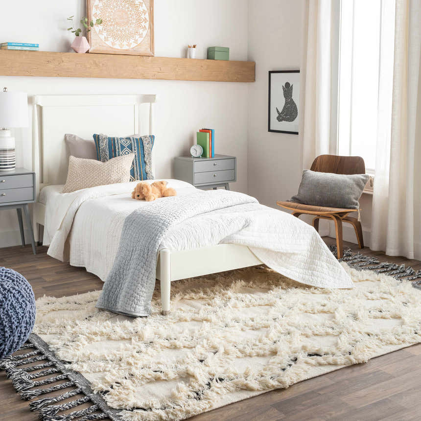 Bedroom scene with a white bed and the cream and black Sangerville Area Rug, a hand woven wool rug with tassels. Roomscene-Rectangle