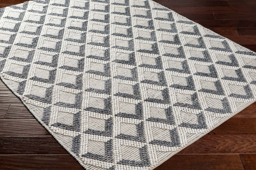 Close-up of the Maik Area Rug. Hand-woven geometric design in dark gray and light gray on a dark wood floor. Corner