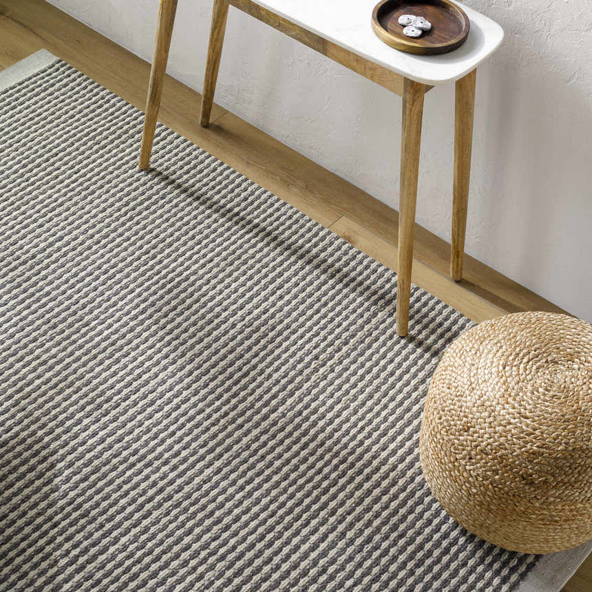 Close-up of the Black Emlen Area Rug showing its woven texture and gray and cream stripes, styled with a side table and pouf. Styleshot
