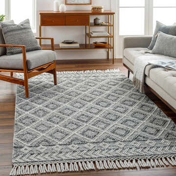 Living room scene with gray sofa and chair, showcasing the dark gray Sook Area Rug with diamond pattern and fringe. Roomscene-Rectangle