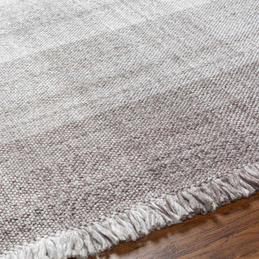 Close-up of the hand-woven Fiora Area Rug showing the gray tones, textured weave, and fringe detail on a wood floor. Texture