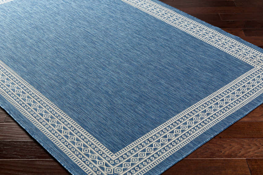 Close-up of the Aleli Blue Area Rug, a contemporary rug with a geometric border on dark wood flooring. Corner