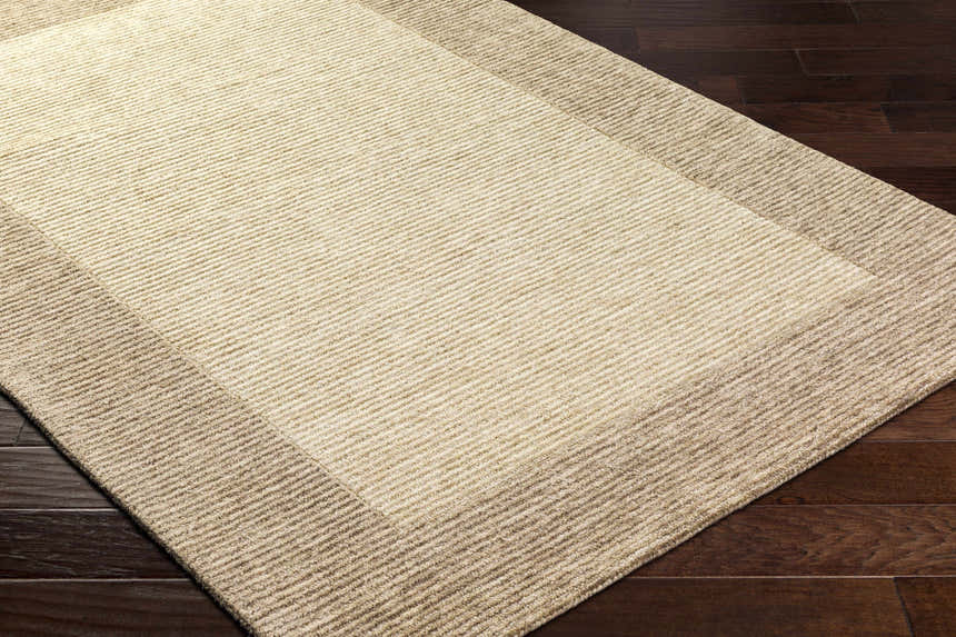 Close up of the hand-tufted Amadi Area Rug in pearl and tan, showing texture and border detail on dark wood floor. Corner
