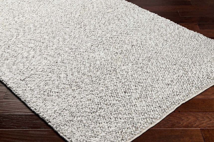 Close-up of the hand woven Chole Area Rug. Neutral gray tones and textured design on dark wood floors. Corner