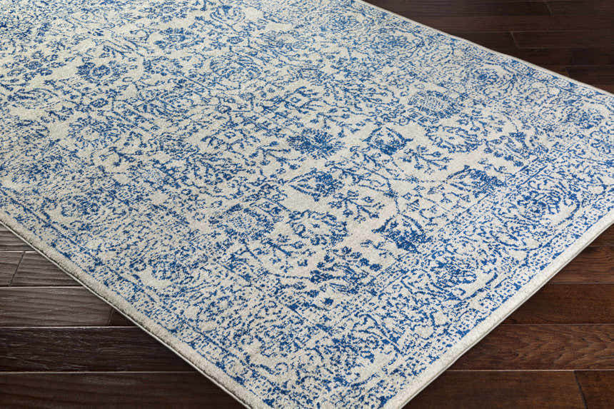 Close-up of the Truxton Area Rug. Light beige rug with a medium blue, vintage-style pattern on dark wood flooring. Corner