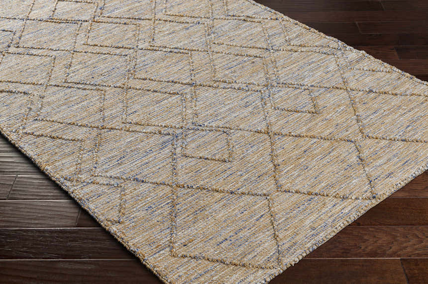 Close-up of the Buse Hand Woven Area Rug. Beige jute rug with a subtle blue geometric diamond pattern on dark brown wood floor. Corner