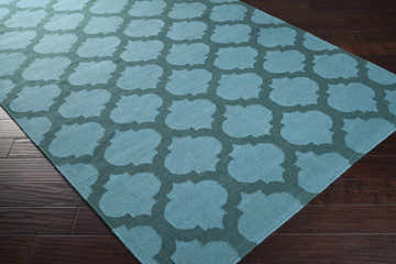 Close-up of the Winside Area Rug, a hand-woven wool rug in teal and dark green quatrefoil pattern on dark wood floors. Corner
