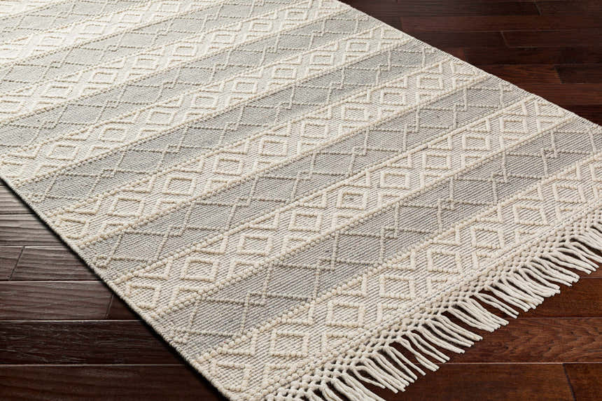 Close-up of Zaza Wool Rug with Tassels. Bone and cloud gray geometric pattern, hand woven texture, and fringe detail. Corner
