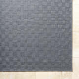 Close-up of the gray Arifah Area Rug showing a woven checkerboard pattern and texture on a light stone tile floor. Corner