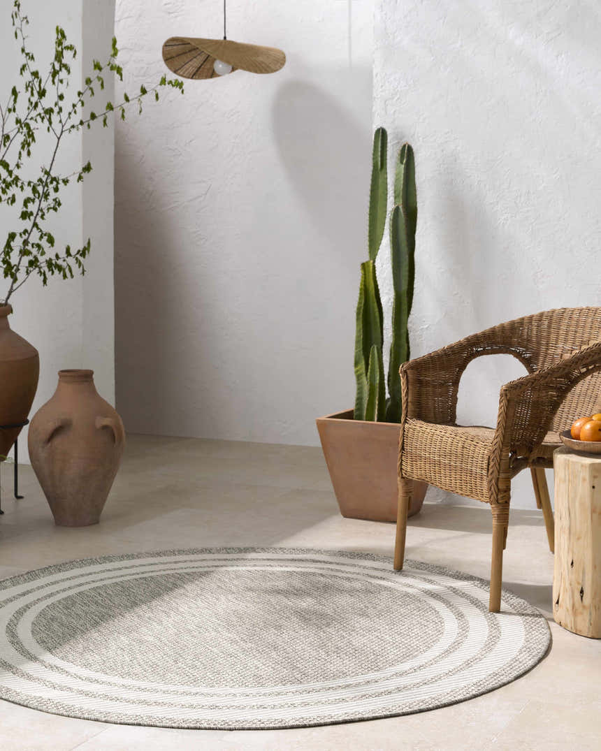 Minimalist patio scene with a round Burradoo Bordered Area Rug in beige and gray under a wicker chair and side table. Roomscene-Round