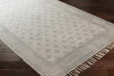 Close-up of the hand-woven Brushton Area Rug. Beige and black geometric pattern with fringe detail on dark wood floor. Corner