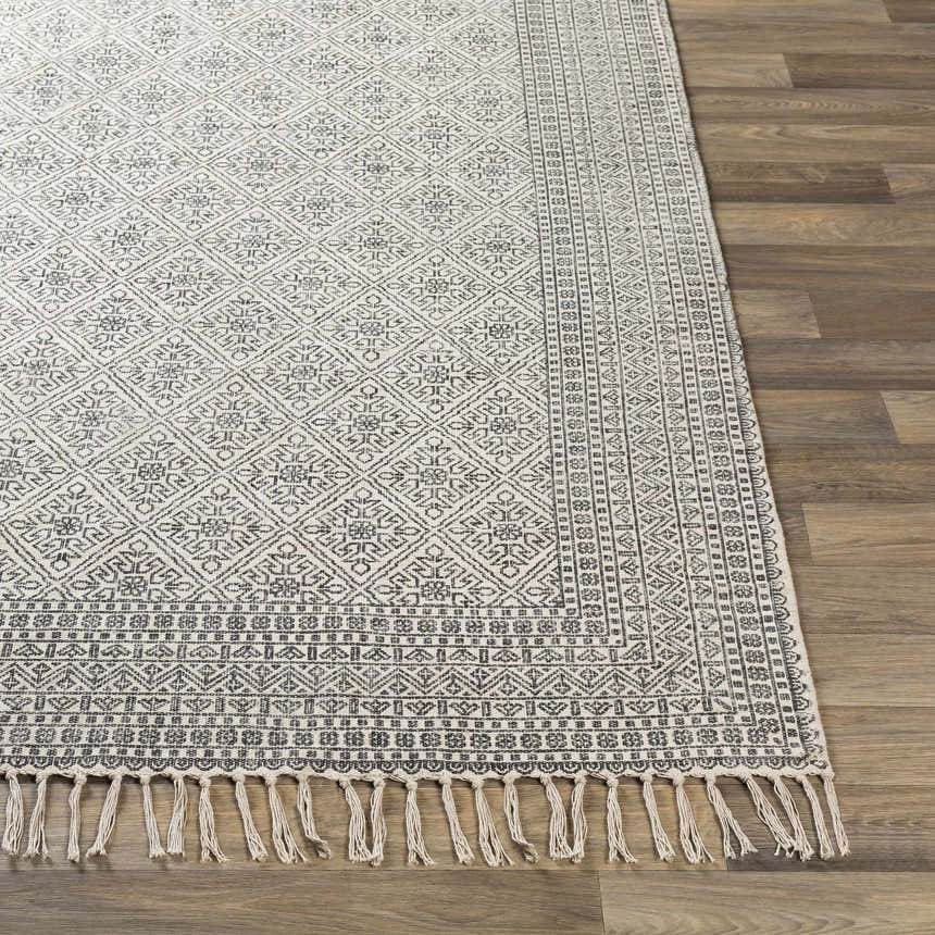 Close-up of the hand woven Brushton Area Rug. Beige cotton rug with black geometric and floral pattern and fringe. Front