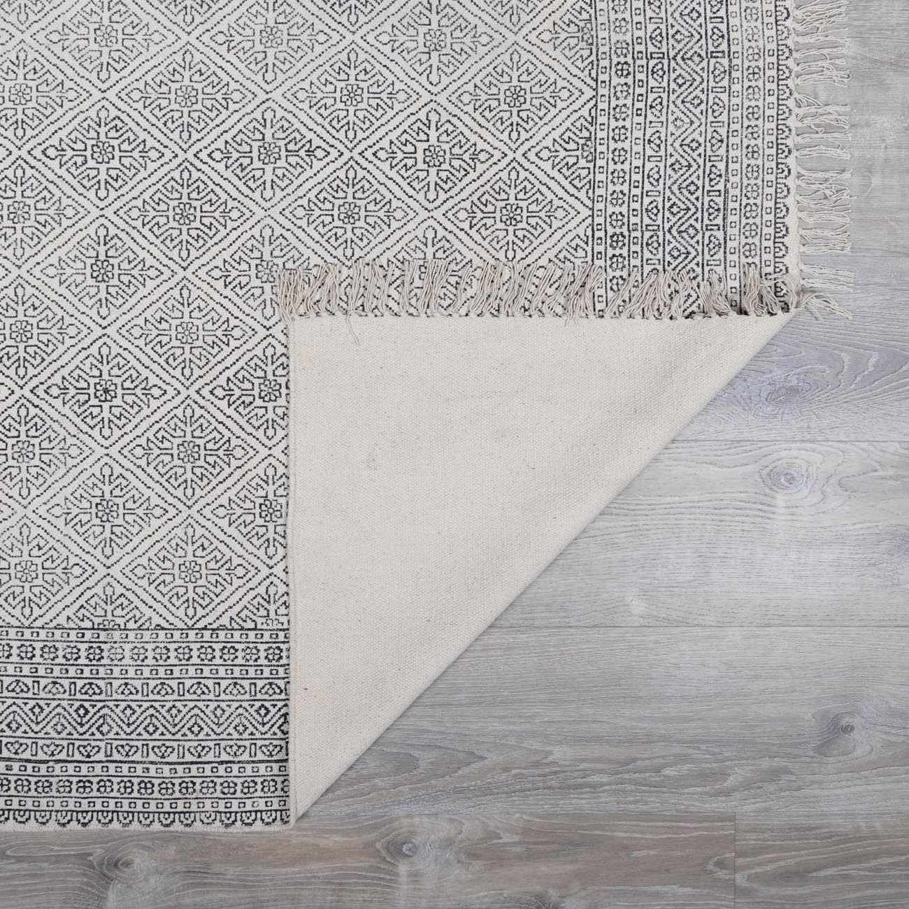 Close-up of Brushton Area Rug with beige cotton backing, black geometric pattern, and fringe on light gray wood floor. Fold