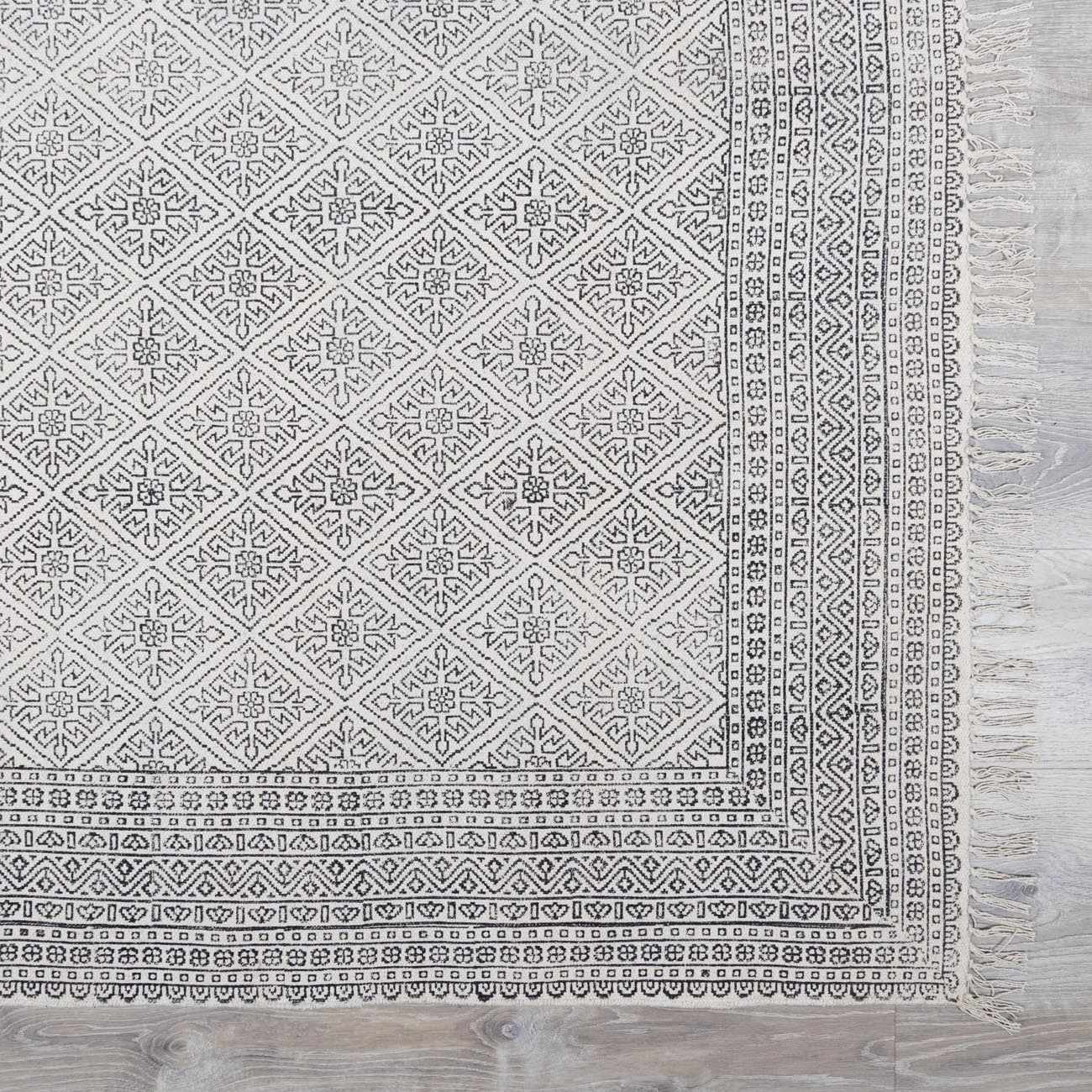 Close-up of the Brushton Area Rug. Beige cotton rug with black geometric diamond and border patterns, and fringe. Swatch