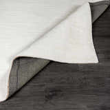 Close-up of the white, hand-loomed Brownsdale Area Rug showing its texture and backing on dark wood floors. Fold