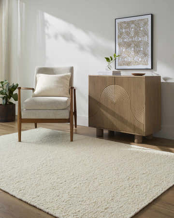 Living room scene with a Bogdan Off-White Wool Rug, gray armchair, and modern wood cabinet in a sunlit, minimalist space. Roomscene-Rectangle