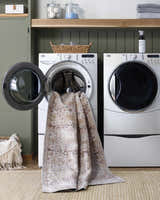 Laundry room scene with Bienna Vintage Flat Pile Washable Rug partially in the washing machine, showing its camel and cream colors. Washing-Machine
