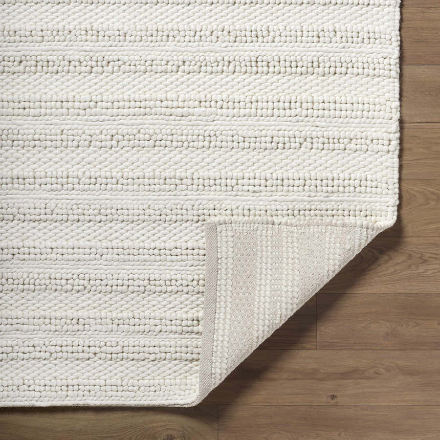 Close-up of the hand-woven Coskun Area Rug. Features a textured, looped pile design in creamy white wool on wood floors. Fold