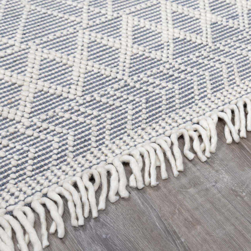 Close-up of the Ramsbury Denim Blue Wool Rug featuring a geometric pattern and ivory fringe detail. Corner