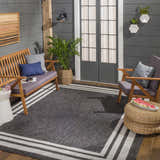 Outdoor patio scene with Coonamble Bordered Antrasit Rug, wood furniture, woven ottoman, and potted plants. Charcoal and cream colors. Roomscene-Rectangle