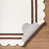 Close-up of the Chogan Ivory Area Rug showing the scalloped edge, brown stripes, and rug backing on wood floor. Fold