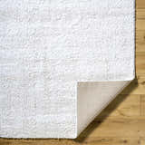 Close-up of the Arlind Area Rug showing its white shag texture and woven backing on a wood floor. Fold