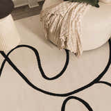 Close-up of the Eilir Modern Boho Rug featuring a bone color and bold, black abstract line design, styled with a round chair. Styleshot