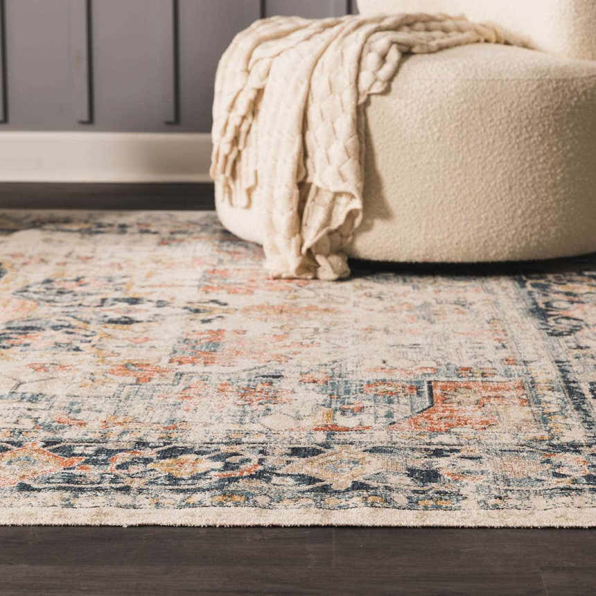 Close-up of the Dorval Traditional Rug in brick red, cream, and navy, styled with a cream ottoman and throw blanket. Styleshot