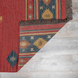 Close-up of the Hedon Wool Area Rug. Brick red rug with southwestern-style geometric patterns in blue, tan, and brown. Fringe detail. Fold