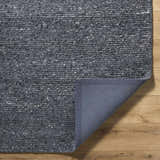 Close-up of the textured Winta Black Wool Area Rug showing gray tones and backing on wood floor. Fold