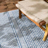 Close-up of the Madras Light Blue Green All Over Performance Area Rug with blue and white geometric patterns, next to a woven chair. Styleshot
