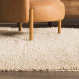 Close-up of the cream Oakridge Area Rug, a hand-woven wool rug with a textured, looped pile, under a tan leather chair. Styleshot